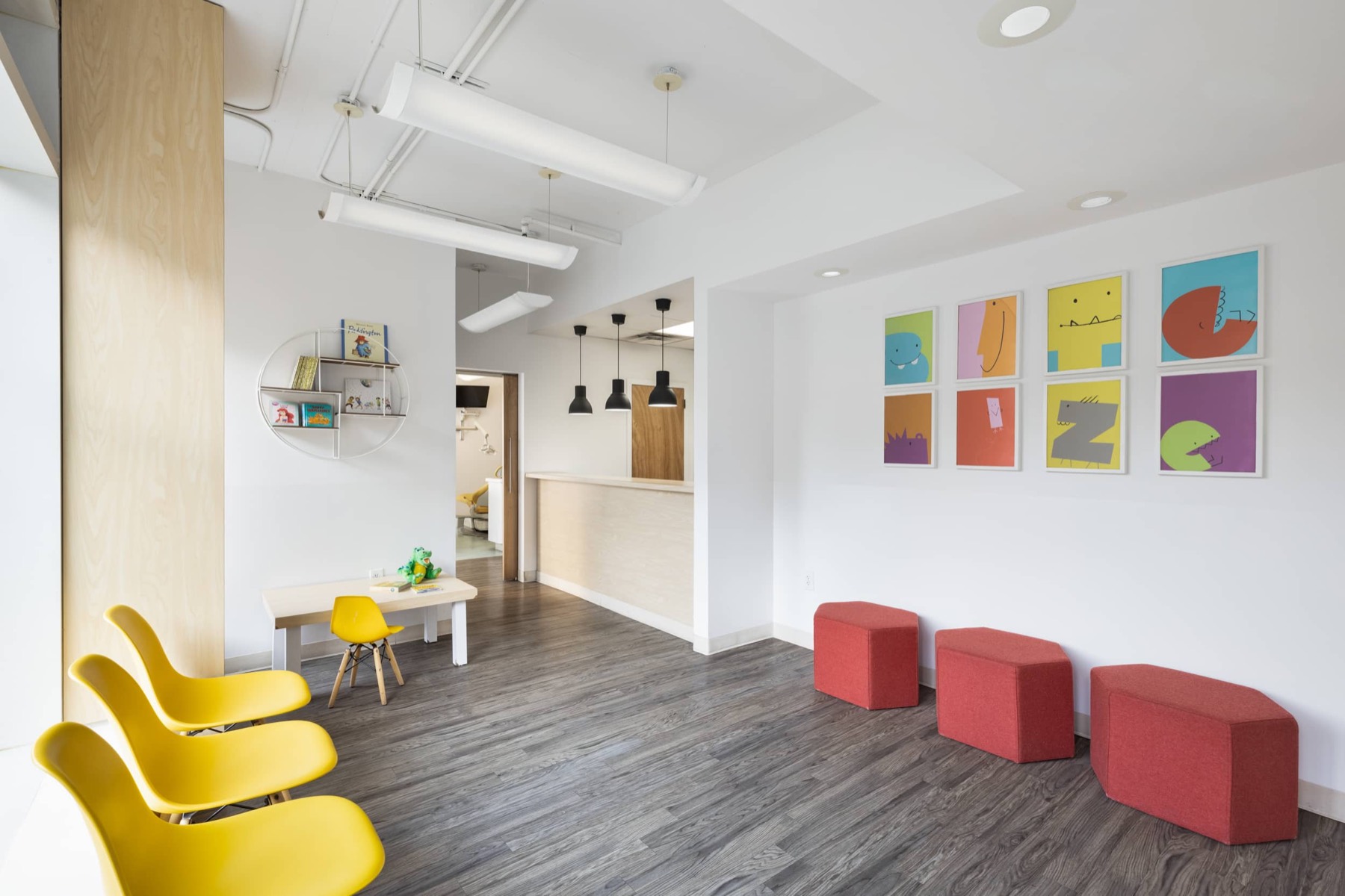 The Bitesize Williamsburg waiting room, yellow shell chairs and coral benches on grey plank flooring with a grid of eight framed monster posters on the white wall