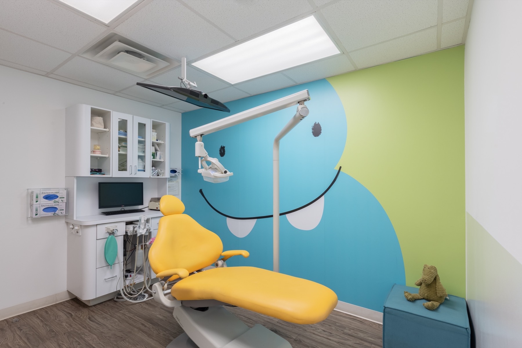 A private treatment room with a whole wall painted as a smiling sky blue monster on a lime green wall, a yellow dental chair and a knitted elephant toy on the bench